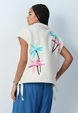 Donna con capelli intrecciati che indossa un gilet senza maniche in pile bianco con ricamo colorato di palme sulla schiena e una gonna blu.