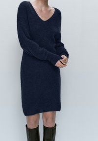 Kvinde iført en langærmet marineblå fuzzy sweaterkjole og sorte knæhøje støvler, der står imod en ensfarvet lys baggrund.