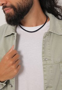 Fekete fonott nyaklánc fehér póló fölött, világoszöld farmerdzsekivel párosítva. A texturált nyaklánc kontrasztot képez a sima anyagrétegekkel.