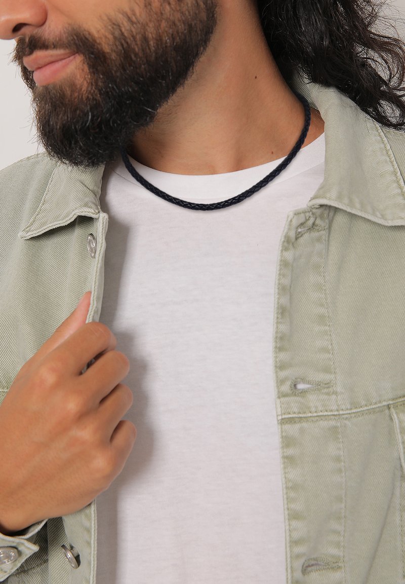 Collana nera intrecciata indossata sopra una maglietta bianca, con una giacca di jeans verde chiaro. La collana strutturata contrasta con gli strati di tessuto lisci.