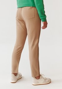Beige slim-fit nadrág sima textúrával, két hátsó zsebbel. Világos színű sportcipőkkel és zöld pulóverrel párosítva.