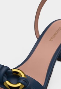 Sandale en cuir bleu à talon moyen, avec une boucle circulaire dorée et une semelle intérieure rose portant le nom de la marque.