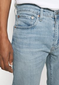 Jean en denim bleu clair avec une texture légèrement délavée. Il présente une taille haute, des poches avant et des accents en métal de ton cuivre.