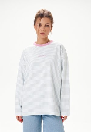 Femme aux cheveux châtain clair attachés, portant une chemise rayée bleu clair à manches longues avec un col rose et un jean bleu clair, expression neutre.