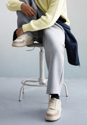 Beige Sportschuhe mit strukturiertem Obermaterial, mehrlagigem Design und Gummisohle, kombiniert mit grauen Jogginghosen und einem hellgelben Strickpullover.