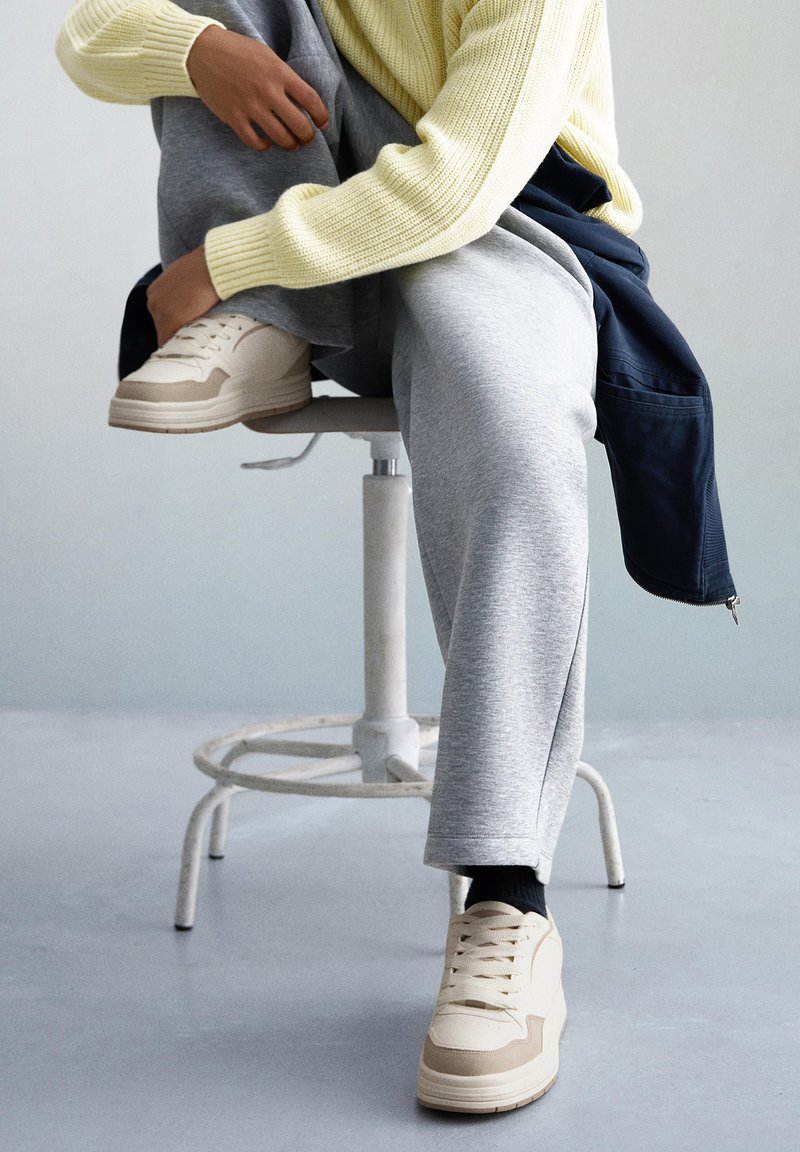 Bež sportske tenisice s teksturiranim gornjim dijelom, slojevitim dizajnom i gumenim potplatom, uparene sa sivim trenirkama i lagano žutim pletenim džemperom.