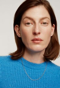 Suéter azul de punto con un patr�ón texturizado, combinado con un delicado collar de oro rosa fino. Un maquillaje neutro complementa el conjunto.