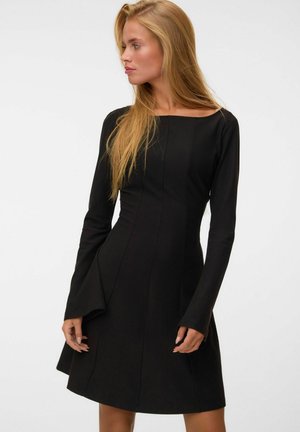 Donna con i capelli biondi lunghi che indossa un abito nero aderente a maniche lunghe, in piedi davanti a uno sfondo bianco uniforme, che guarda di lato.