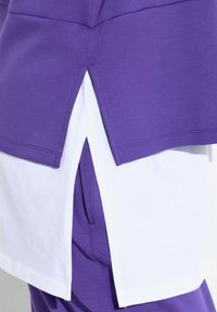 Top a strati viola e bianco con design a fessura, caratterizzato da un sottostante bianco visibile e una finitura morbida e texturizzata.