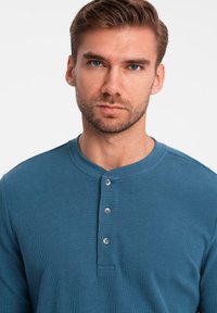 Mann mit kurzen braunen Haaren und Bart, der ein blaues langärmliges Henley-Shirt mit drei Knöpfen trägt und mit dem Gesicht nach vorne vor einem einfachen hellen Hintergrund steht.