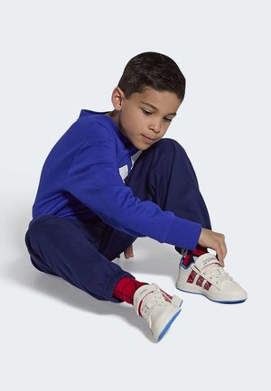 Baskets blanches avec des accents rouges et bleus, des sangles texturées et un motif distinctif sur le côté. L'enfant porte un t-shirt bleu et un pantalon marine.