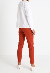 Woman wearing a white long-sleeve button-back shirt, rust-colored slim-fit pants, and white slip-on sneakers, standing against a white background.