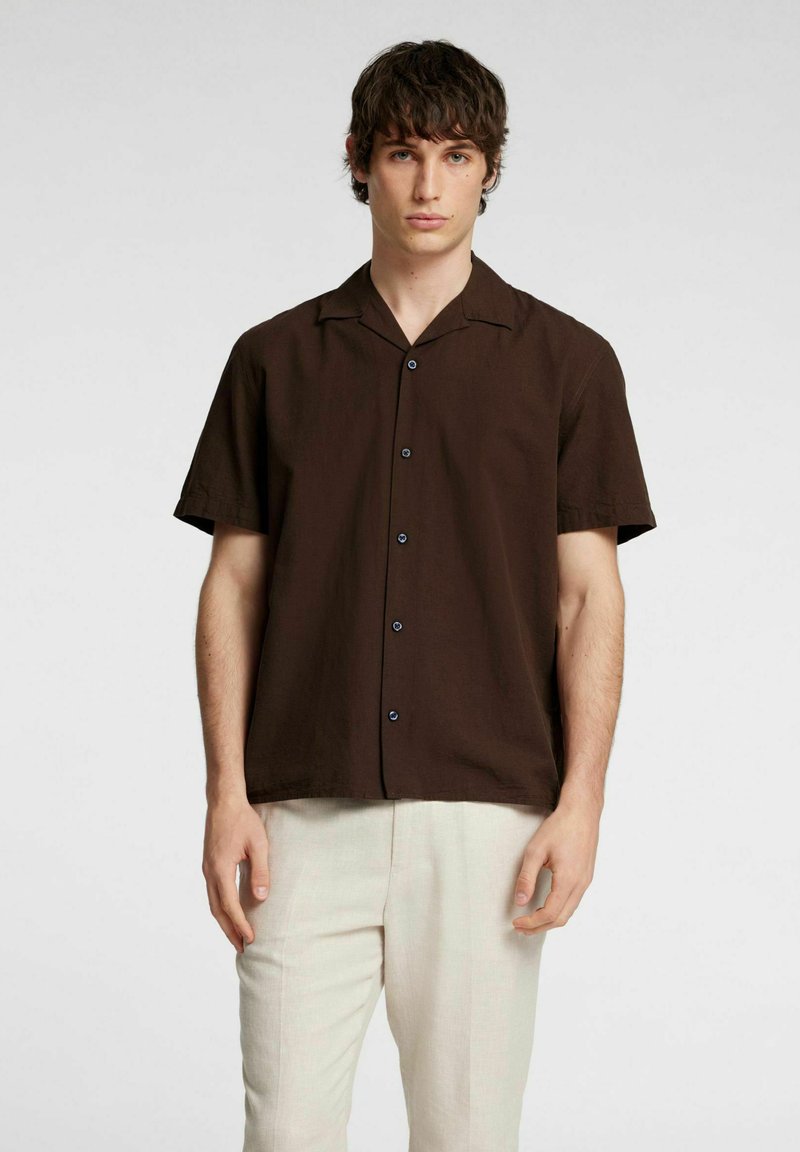 Camicetta marrone a maniche corte con colletto a punta, caratterizzata da bottoni neri e una vestibilità rilassata. Abbinata a pantaloni chiari e testurizzati.