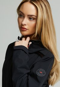 Zwarte waterdichte jas heeft een verstelbare capuchon met trekkoorden, rode stiksels en een ronde patch met de tekst "Zavetti Canada" op de mouw.