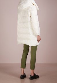 Vit puffig kappa med huva och kviltat mönster, långa ärmar och midje-lång längd, tillsammans med olivgröna tights och svarta skor.