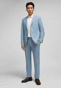 Lichtblauw pak met een gestructureerd patroon, uitgerust met een jasje met twee knopen en slim-fit broek. Gecombineerd met een witte blouse met kraag en bruine loafers.