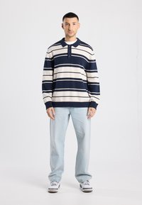 Marineblauw en crème gestreepte gebreide polotrui met drieknopenbies, geribbelde manchetten en zoom. Gecombineerd met lichtblauwe, relaxed-fit jeans en sneakers.