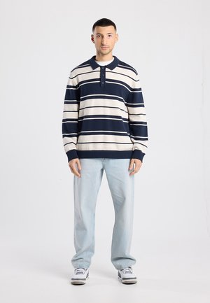 Marineblauw en crème gestreepte gebreide polotrui met drieknopenbies, geribbelde manchetten en zoom. Gecombineerd met lichtblauwe, relaxed-fit jeans en sneakers.
