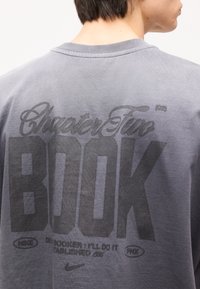 Άτομο που φοράει γκρι πουκάμισο με μεγάλο μαύρο κείμενο που γράφει «Chapter Two BOOK» και μικρότερο κείμενο μαζί με λογότυπα της Nike στην πλάτη.