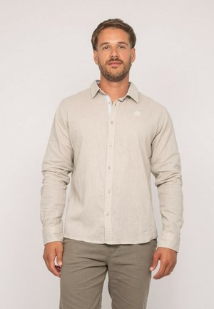 Homme barbu portant une chemise beige à manches longues boutonnée et un pantalon assorti, debout devant un fond clair uni, faisant face à l'avant.