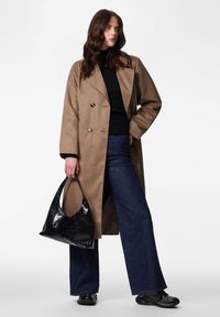 Hosszú, bézs trench coat dupla soros gombokkal, fekete pulóverrel és bővülő szárú farmernadrággal párosítva. Egy fényes fekete hobo táskát tart a kezében.