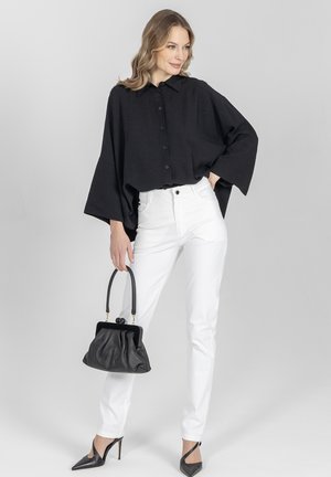 Kobieta ubrana w luźną czarną koszulę na guziki, białe spodnie, czarne szpilki, trzyma czarną torebkę, stoi na jednolitym szarym tle.