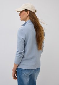 Pull-over en bleu clair avec col et texture côtelée. Associé à un jean en denim bleu clair et une casquette beige. Vue de dos.