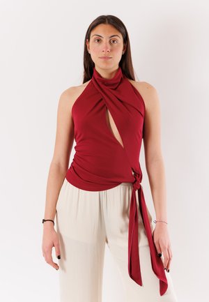 Donna con capelli lunghi e scuri che indossa un top rosso senza maniche con arricciatura frontale e laccio, abbinato a pantaloni bianchi larghi.