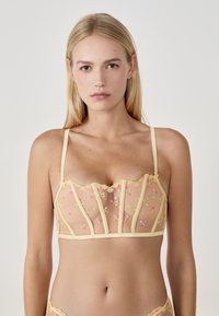 Gelbes transparentes Bralette mit Blumenstickereien, verstellbaren Trägern, gezackten Rändern und einem strukturierten Design mit vertikalen Nähten.