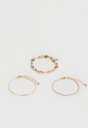 Τρία λεπτά χρυσά βραχιόλια με ρυθμιζόμενα κούμπωματα αλυσίδας, το ένα με οβάλ πολύτιμους λίθους, τοποθετημένα σε απλό λευκό φόντο.