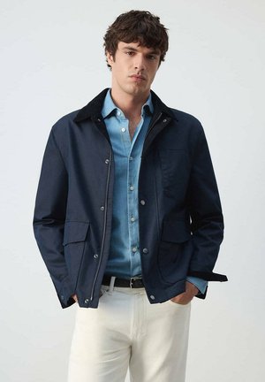 Joven con chaqueta azul marino sobre una camisa azul de botones y pantalones blancos con las manos en los bolsillos de la chaqueta sobre un fondo liso.