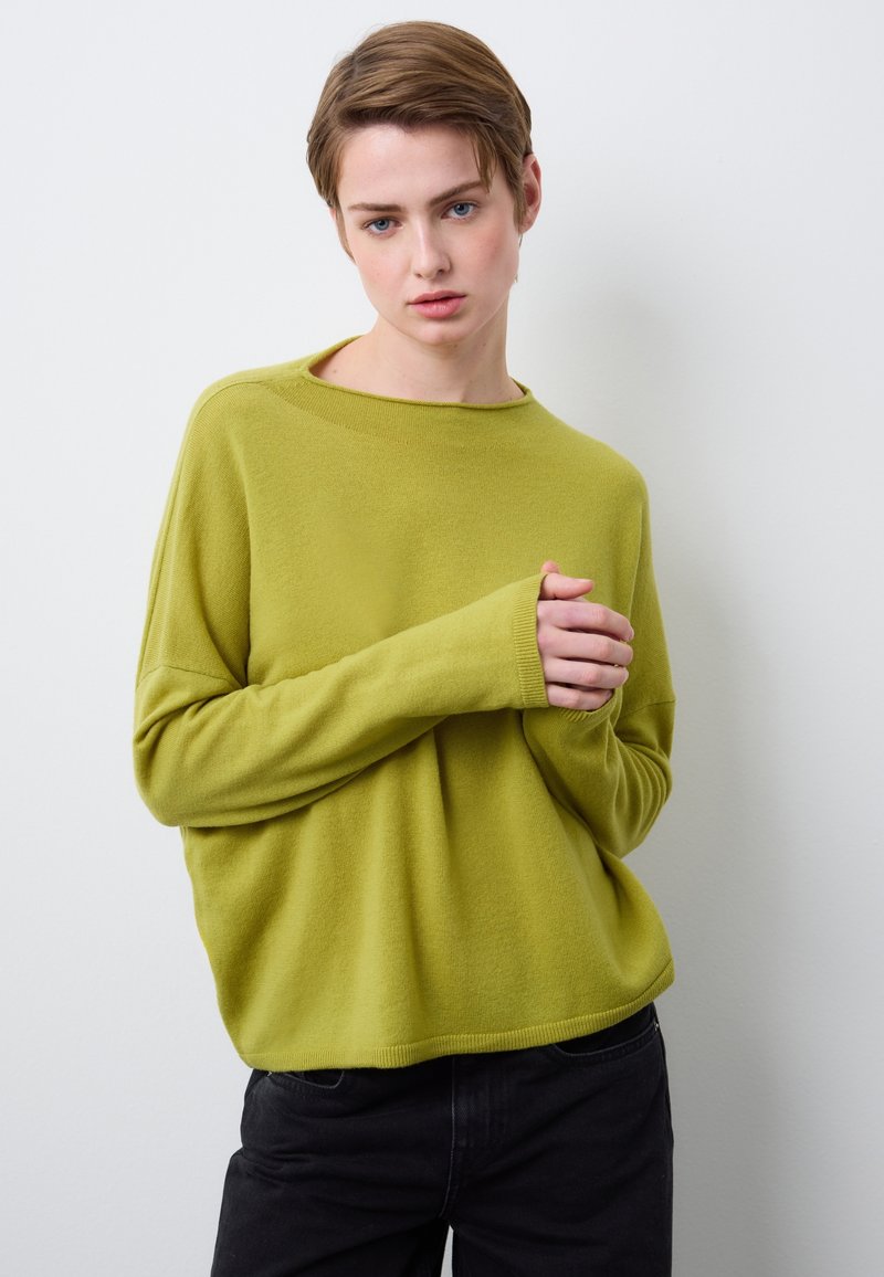 Grüner Strickpullover mit langen Ärmeln, weitem Ausschnitt und gerippten Bündchen, kombiniert mit dunklen Jeans vor schlichter Hintergrund.
