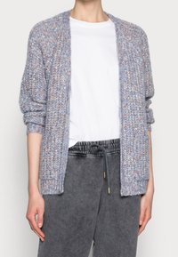 Světle modrý pletený cardigan s tlustou strukturou, otevřeným předním dílem a žebrovanými manžetami. V kombinaci s bílým tričkem a šedými kalhotami se šňůrkou.