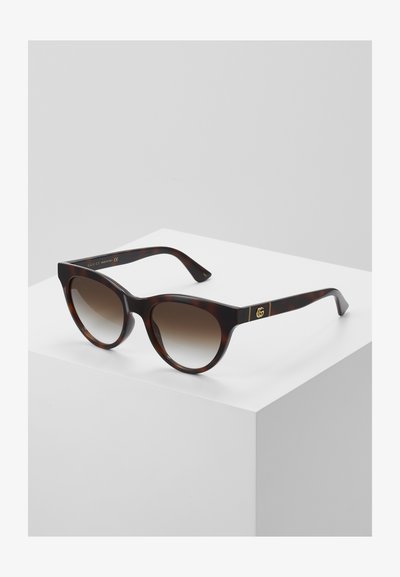 Tortoiseshell sunglasses with a cat-eye shape, gradient brown lenses, gold logo accents on the arms, sleek, glossy finish.