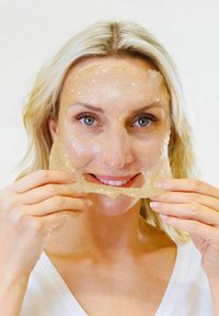 Guld glimmer gelmaske holdt af en person. Masken har en glat, gennemsigtig tekstur og er med udskæringer til ansigtstræk.