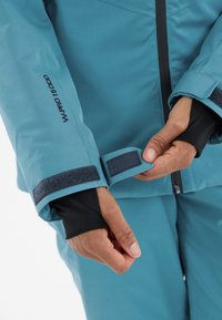 Türkisfarbene wasserdichte Jacke mit schwarzen Akzenten, ausgestattet mit Klettverschluss am Ärmelbund und einem weichen Innenfutter für Wärme und Komfort.