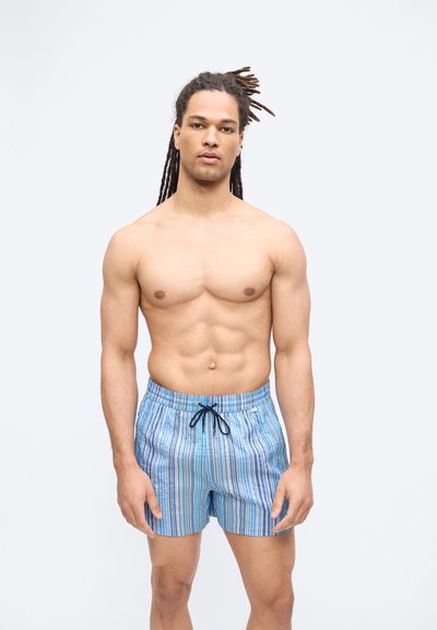 Άνδρας με μακριές πλεξούδες ντυμένος με μπλε ριγωτό μαγιό, στέκεται μπροστά σε απλό λευκό φόντο.