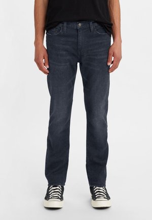 Homme portant un jean droit bleu foncé et des baskets montantes noires, debout devant un fond clair uni.
