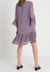 Robe florale avec un motif violet et rose, manches évasées trois-quarts et un ourlet à volants. Confectionnée en tissu léger et texturé.