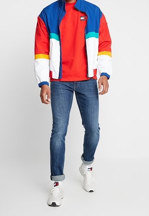 Chaqueta cortavientos colorida en azul, rojo, blanco y amarillo con cierre frontal; llevada sobre una camiseta roja, combinada con vaqueros azules y zapatillas blancas.