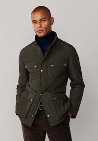 Kvadrični, maslinasto zeleni jakna sa dugmadi na prekidač, dva prednja džepa i visokim ovratnikom, nosi se preko mornarsko plave turtleneck majice.