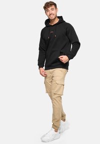 Sort hættejakke med en frontlomme, snørelukning og broderet logo. Parret med beige cargobukser og hvide sneakers.