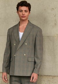 Šedý kostkovaný dvouřadý blazer s jemnými oranžovými akcenty, klopovými límci a dvěma předními kapsami. Nesený přes bílou košili.