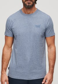 T-shirt à manches courtes bleu chiné en tissu doux, avec un petit logo d'un bleu légèrement plus foncé sur la poitrine gauche.