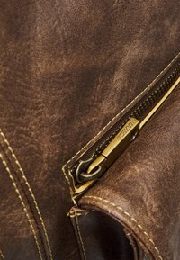 Sac en cuir marron avec une fermeture éclair en laiton visible, des coutures jaunes, une surface texturée et un détail de poche, mettant en valeur le matériau et le savoir-faire.