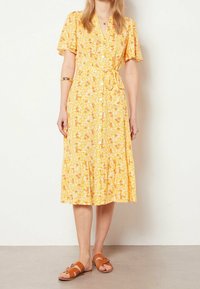 Abito midi giallo a fiori con maniche corte a volant, chiusura con bottoni e cintura. Abbinato a sandali scivolati marroni.