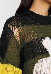 Pull tricoté avec un tissage lâche et ajouré en rayures vertes et noires, orné d'un motif floral blanc, souligné par un col côtelé et des accents jaunes.