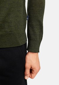 Olivgrüner gestrickter Pullover mit strukturiertem Finish, gerippten Bündchen und einem Markenetikett. Über schwarzen Stoffhosen getragen, zeigt eine Hand, die die Seite greift.