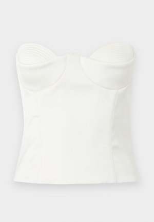 Ženska z dolgimi temnimi lasmi, ki nosi bel oprijet korzet brez naramnic in ujemajočo dolgo krilo, stoji z eno roko na boku na enostavni podlagi.