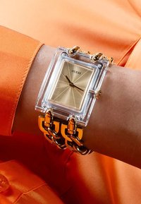 Klar rektangulær ur med en guld skive, sorte visere og en kraftig guldkædearmbånd, båret på et håndled mod en orange stofbaggrund.